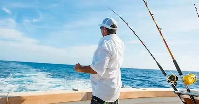 Man on boat fishing in open sea under clear sky with two fishing rods.